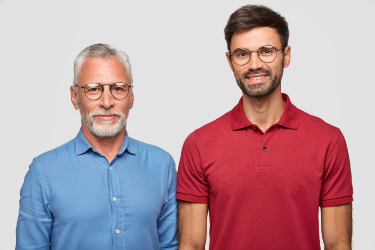 mature-grey-haired-man-and-his-adult-son-stand-against-white-wall-have-pleased-expressions-after-meeting-wear-round-spectacles-being-one-friendly-family-people-and-generation-concept