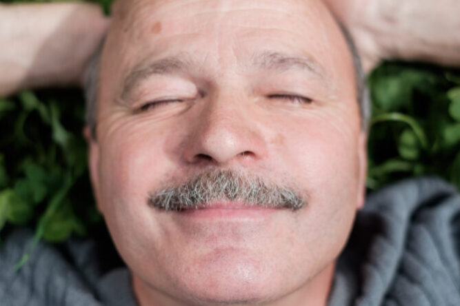 Mature Happy Man Lying On Green Grass. He smiles and closes eyes.